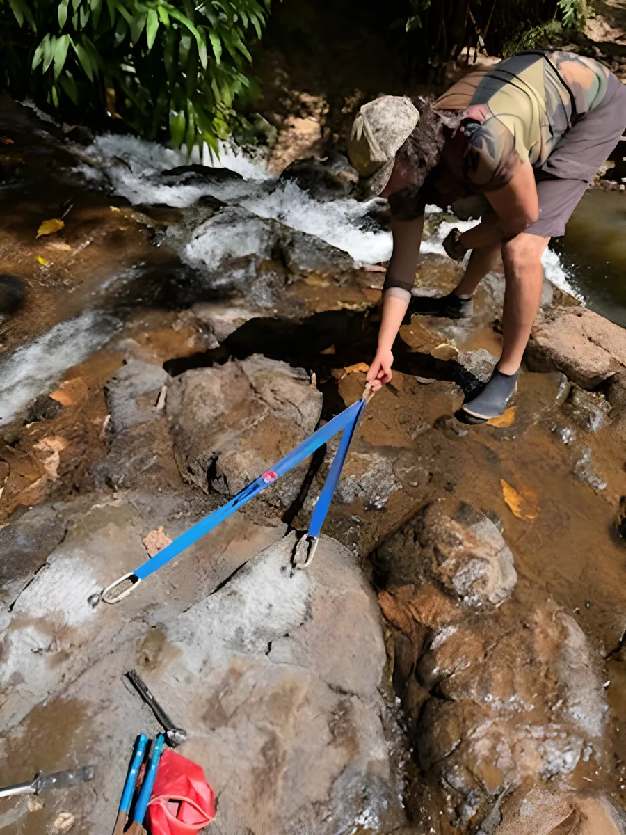 A guide in a camo shirt rigging a blue sling to a two-bolt anchor on wet rock in a stream