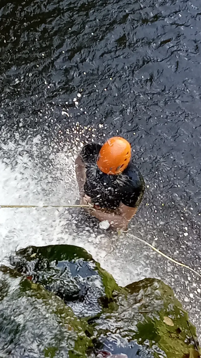 A rappeller in an orange helmet in the spray at the bottom of a waterfall