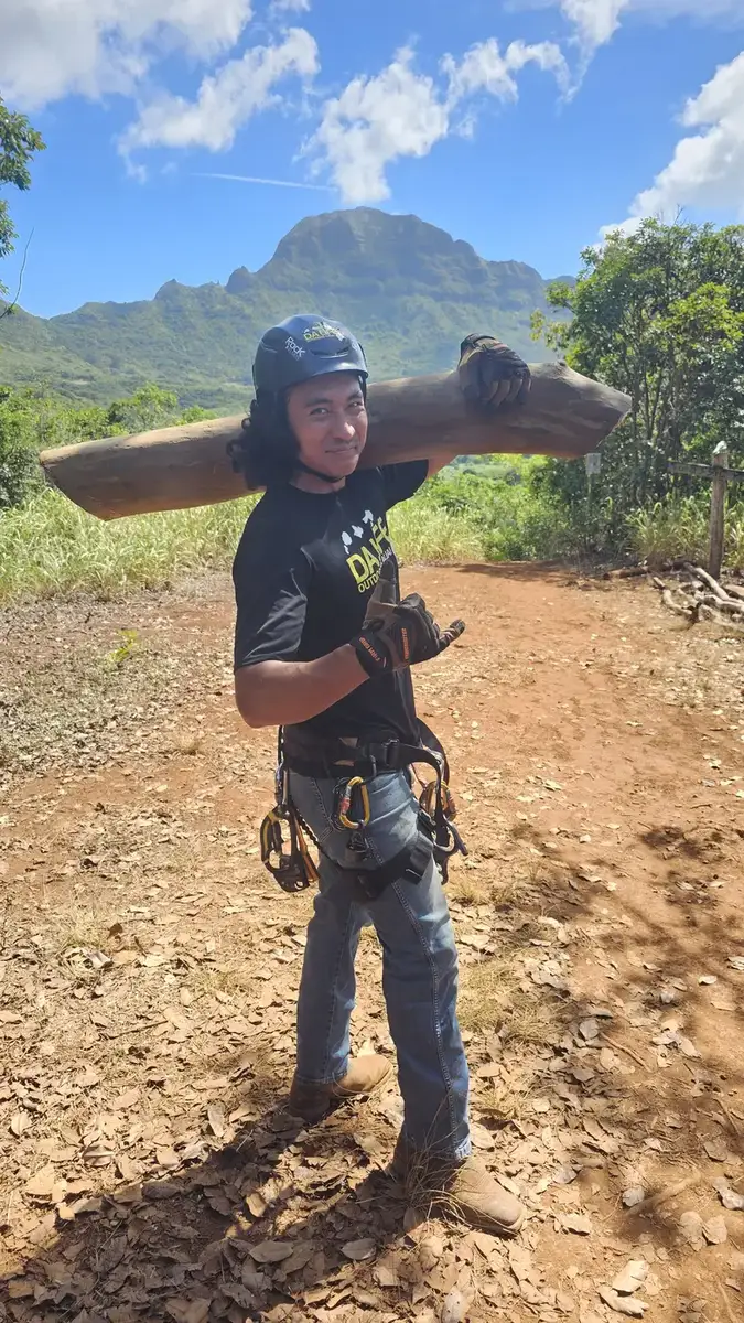 A crew member in a helmet and harness carrying a heavy timber on his shoulder, with mountains behind